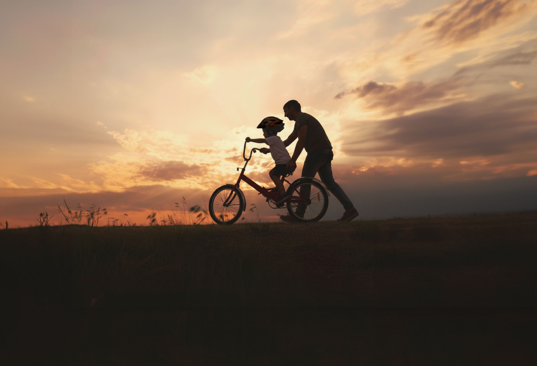 Silhouette d’un adulte aidant un enfant à faire du vélo au coucher du soleil.